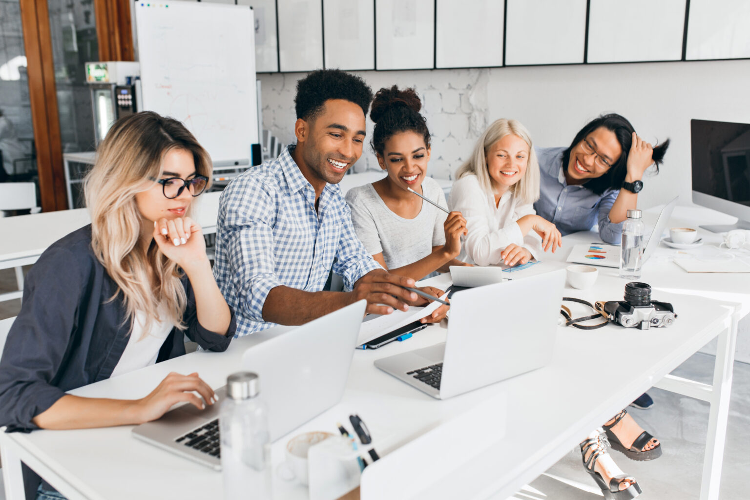 Smiling african student pointing with pencil at laptop screen. Concentrated blonde girl in glasses propping chin with hand while working with computer in office.